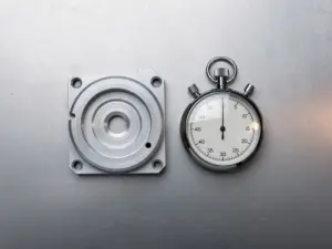 CNC-gefrästes Metallteil mit sichtbaren Bearbeitungsrillen neben einer analogen Stoppuhr auf Stahloberfläche.