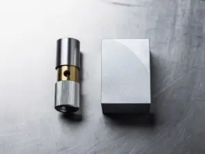 Präzisionsgefräster Aluminiumblock und Metallzylinder mit Messingakzent auf Stahloberfläche, feine Bearbeitungsspuren sichtbar.