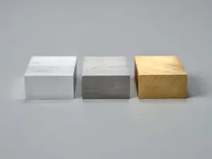 Drei präzisionsgefräste Metallproben aus Aluminium, Stahl und Messing nebeneinander auf grauer Werkstattoberfläche mit sichtbaren Bearbeitungsspuren.