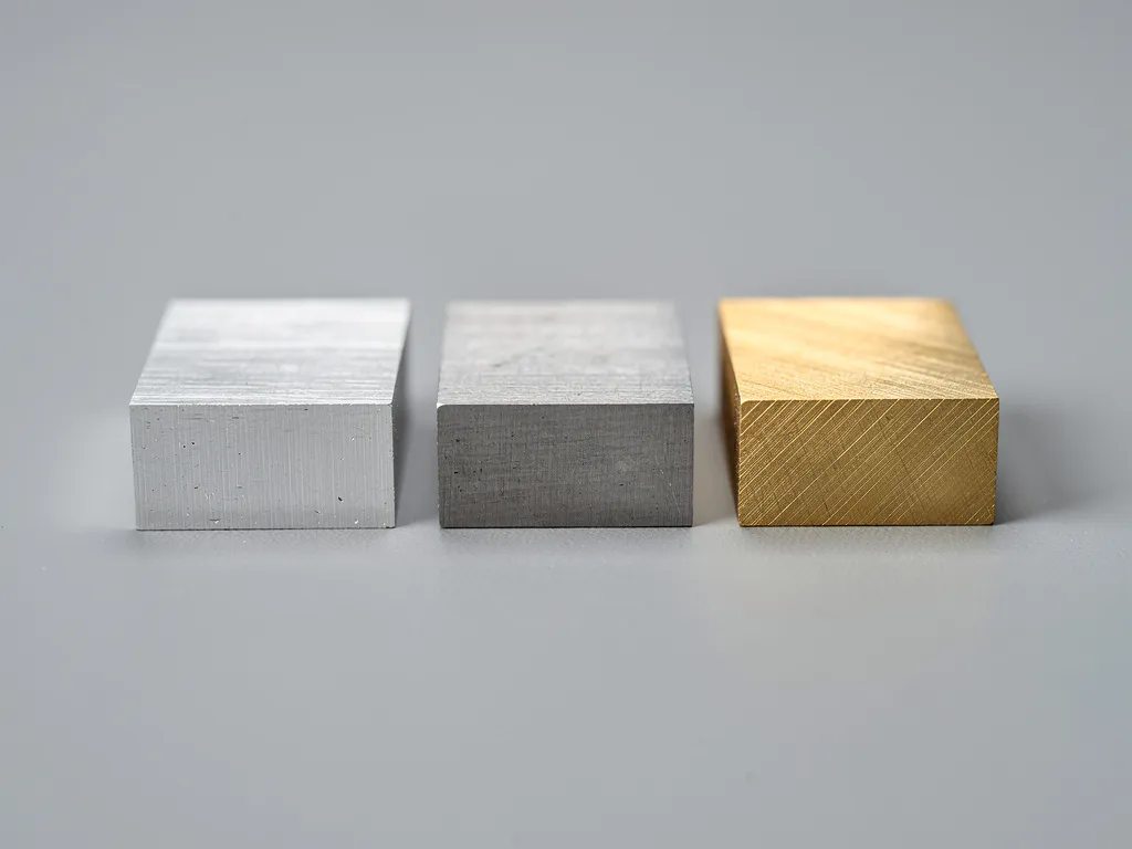 Drei präzisionsgefräste Metallproben aus Aluminium, Stahl und Messing nebeneinander auf grauer Werkstattoberfläche mit sichtbaren Bearbeitungsspuren.