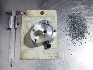 Präzisionsgefrästes Metallteil auf verwittertem Werkstattschein neben Messschieber und Stahlspänen auf Aluminiumoberfläche.