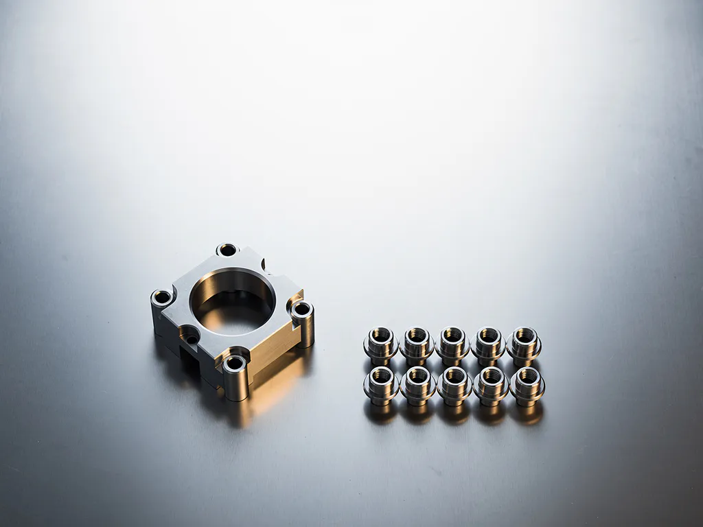 Präzisionsgefertigtes Metallteil neben einer Gruppe identischer Komponenten auf gebürstetem Stahl, Draufsicht mit weichem Industrielicht.