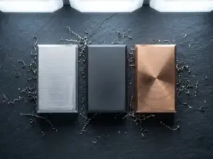 Drei präzisionsgefertigte Metallproben aus Aluminium, Stahl und Bronze auf grauer Werkstattoberfläche mit Metallspänen.