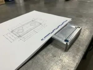 Technische Zeichnung auf mattem Papier neben einem gefrästen Aluminiumteil mit Werkzeugspuren auf einer Stahloberfläche.