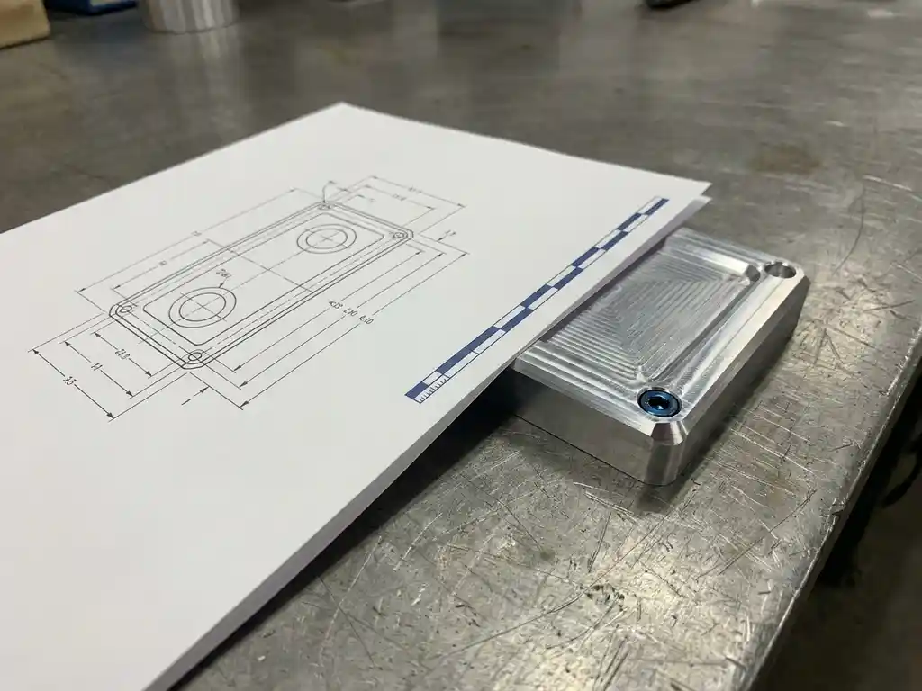 Technische Zeichnung auf mattem Papier neben einem gefrästen Aluminiumteil mit Werkzeugspuren auf einer Stahloberfläche.